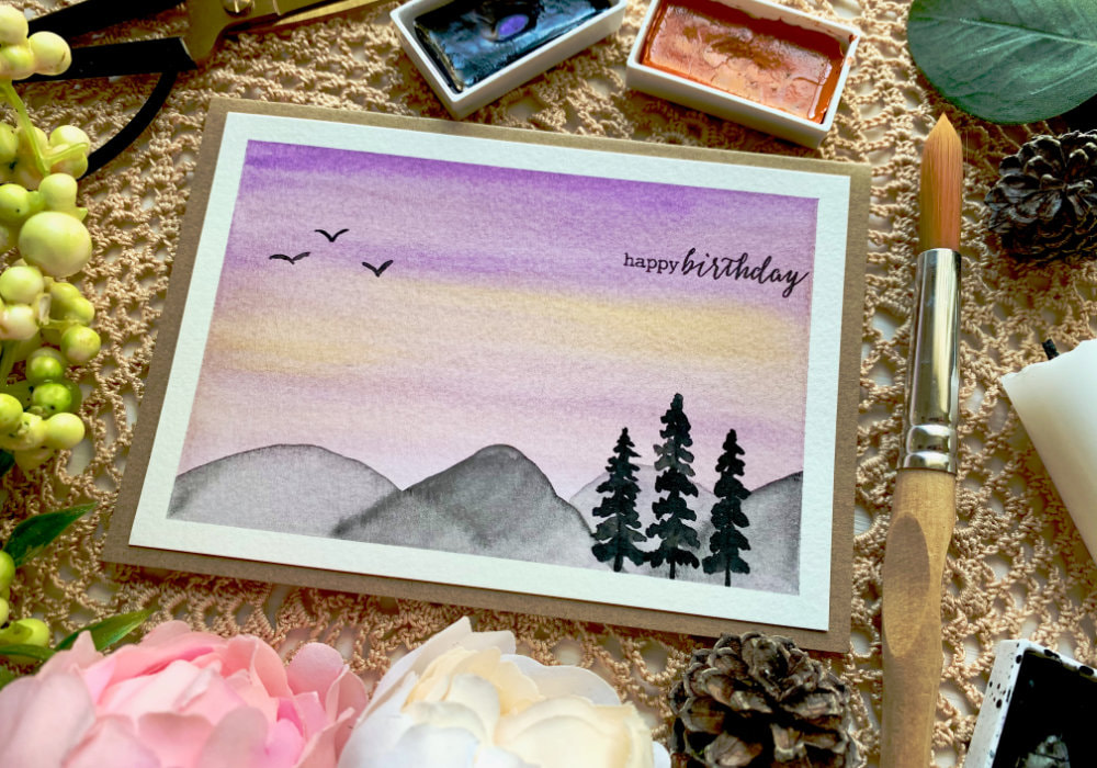 Handmade card with watercolour landscape with purple sky and grey mountains with trees.