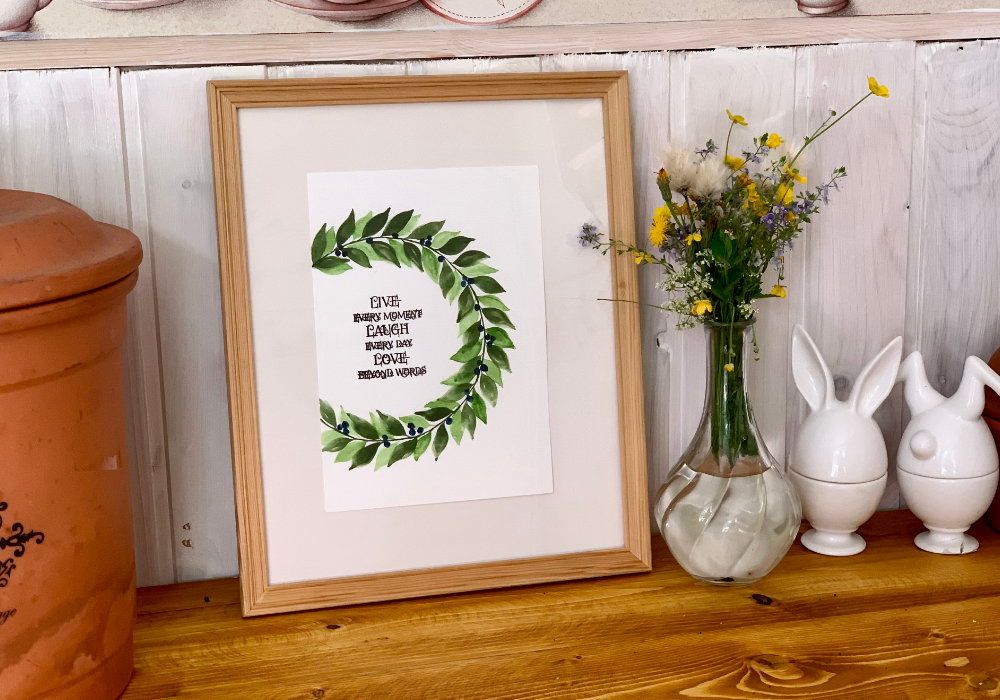 Watercolour painting of a wreath in two shades of green and blue berries. One of the paintings is made into a greeting card with a stamped greeting in the middle saying Have A Lovely Day. The other paining is placed into a picture frame and in the middle has a stamped quote saying  Live Every Moment, Laugh Every Day, Love Beyond Words.