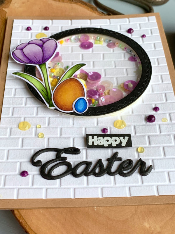 Handmade greeting card for Easter with a circle shaker element filled with sequins and beads, crocus flower and eggs on top of the shaker, a white background panel with a brick pattern created using an embossing folder and the word Easter die-cut from a black card stock and the word happy stamped and heat embossed in white and cut into a banner. All adhered on top of a card base made out of a craft card stock.