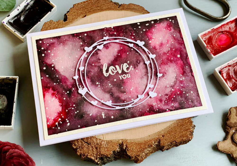 Handmade card for Valentine's day or anniversary with a pink watercolour background of a galaxy, greeting that says Love You stamped and heat embossed in the middle and white die cut with heart wreath attached in the middle of the card.
