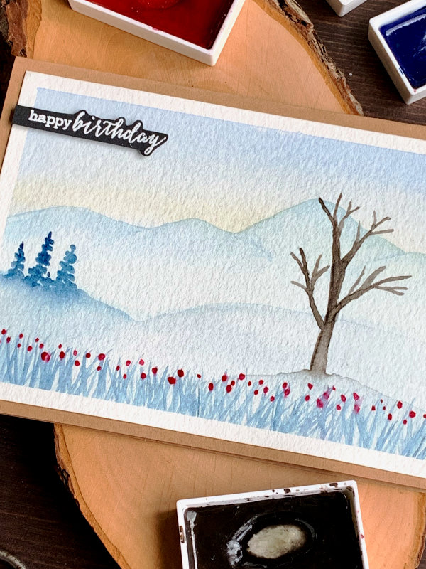 Handmade card with a winter watercolour landscape. With snowy mountains in the background, small pine trees in the middle and a big tree, without leaves in the front.