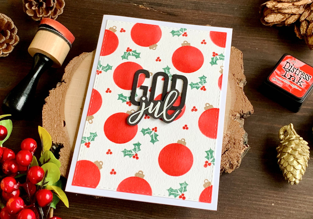 Handmade Christmas card with baubles created with Distress inks and a (DIY) circle stencil. Background filled with small red baubles with golden tops with loops. In between are stamped holly berry and leaves. The greeting says God Jul and is created with individual letter stamps and dies.