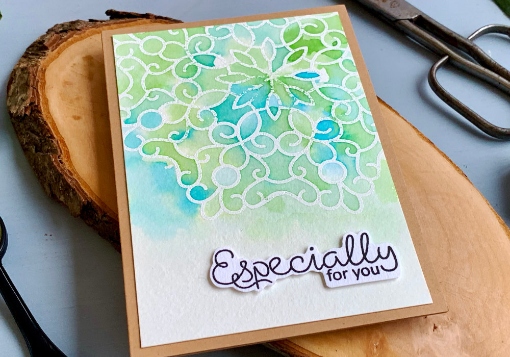 Blue and green Distress ink watercolour colour combination using the inks Salty Ocean and Mowed Lawn. With a big mandala stamped and heat embossed and watercoloured with the Distress inks creating very light background. 